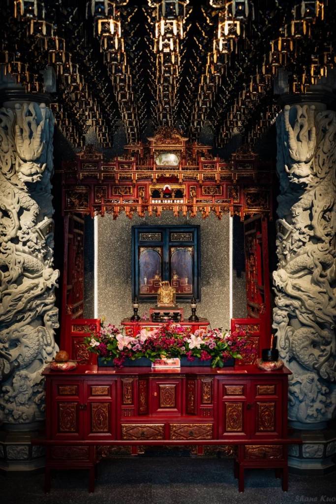 一個裝飾華麗的寺廟內部，中央有供桌和花卉，背景是精美的雕刻和神像。