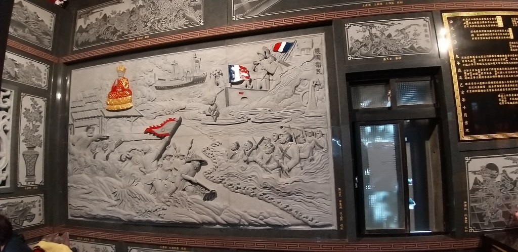 一面雕刻精美的牆壁藝術，描繪了船隻、士兵和歷史場景，配有法國國旗和金色王冠的裝飾。